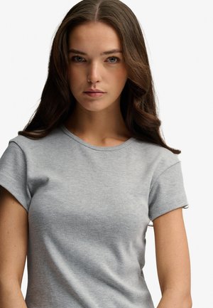 Młoda kobieta z długimi brązowymi włosami, ubrana w dopasowany jasnoszary t-shirt z krótkim rękawem, patrząca przed siebie o neutralnym wyrazie twarzy.