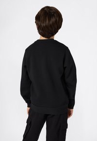 Niño con cabello corto castaño, vistiendo un sudadera negra de manga larga y pantalones cargo negros, de pie mirando hacia atrás sobre un fondo blanco.