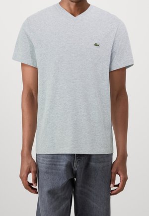 Homme portant un T-shirt gris clair à col en V avec un logo de crocodile vert sur la poitrine et un jean gris foncé, debout devant un fond clair uni.