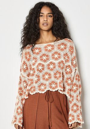 Young woman with long curly hair wears a loose, crochet-patterned top in cream and rust colors with rust-colored high-waisted pants.