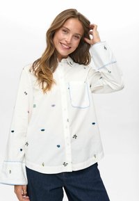 Glunderende vrouw met lang haar, gekleed in een witte overhemd met kleurrijke, kleine geborduurde ontwerpen en donkerblauwe spijkerbroek, terwijl ze in haar haar raakt.