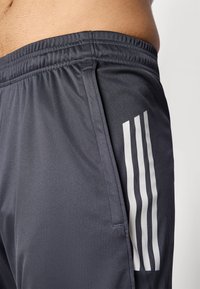 Pantaloni scurți atletici gri, cu talie elastică, având trei dungi albe pe partea stângă și un material neted, texturat.