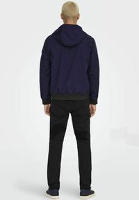 Giacca a cappuccio blu scuro con orlo a costine, abbinata a jeans neri slim-fit. Le scarpe sono scure con suola bianca. La vista posteriore mette in evidenza i dettagli di design.