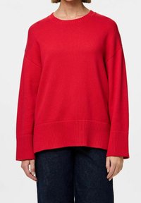 Czerwony sweter z dzianiny z okrągłym dekoltem, opadającymi ramionami oraz ściągaczami u mankietów i na dole. Miękka faktura i luźny krój. Połączony z ciemnymi dżinsami.