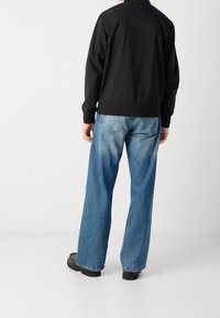 Schwarze Bomberjacke mit gerippten Bündchen, kombiniert mit hellblauen Jeans mit weitem Bein und schwarzen klobigen Stiefeln, von hinten gesehen.