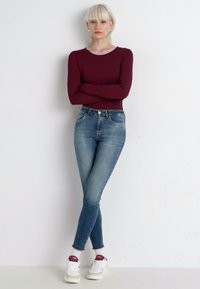 Burgunderfarget langermet skjorte og lys blå skinny jeans, kombinert med hvite joggesko med røde detaljer. Modellen står med armene i kors.