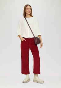 Haut blanc texturé avec des manches larges, associé à un pantalon en velours côtelé rouge taille haute et des bottines beige. Petit sac en bandoulière bordeaux.
