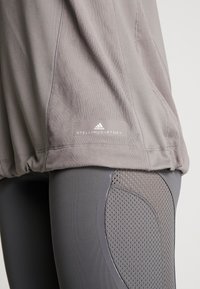 Zbliżenie na szare legginsy sportowe z panelami z siateczki oraz taupe'ową bluzkę o fakturze siateczki z metką logo Adidas by Stella McCartney przy krawędzi.