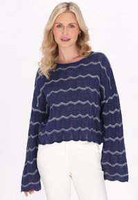 Marineblauer, gestrickter, kürzerer Pullover mit wellenförmigen silbernen Streifen, weiten Ärmeln und einer strukturierten Oberfläche. Kombiniert mit hellen Hosen.