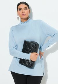 Himmelblaues Langarmoberteil mit Rüsche am hohen Kragen und Knopfakzenten an den Manschetten, kombiniert mit einer schwarzen gepolsterten Clutch.