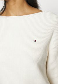 Kermanvärinen ribbikudottu neulepaita, jossa on leveä kaula-aukko ja Tommy Hilfiger -logo punaisena, valkoisena ja sinisenä rinnassa.