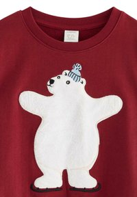 Roter Kapuzenpullover mit einem plüschigen, weißen Eisbär-Design. Der Bär trägt eine hellblaue Strickmütze und steht auf schwarzen Schlittschuhen.