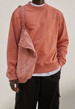 Personne portant un sweat-shirt corail, un pantalon cargo noir et portant un sac en tissu corail assorti sur son épaule.