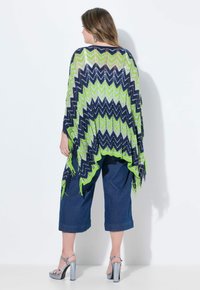 Stickad poncho i marinblått, grönt och vitt zigzag-mönster med fransad kant, bärs över mörk denim culotter, i kombination med silverhöga sandaler.