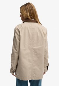 Mujer con cabello rubio que lleva una chaqueta beige con cuello marrón, vista desde atrás, combinada con jeans azules sobre un fondo blanco.