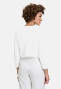 Biały krótki sweter wykonany z puszystego materiału, z rękawami o długości trzy czwarte i okrągłym dekoltem. Nosi się go na białych dolnych częściach odzieży.