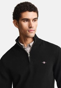 Junger Mann mit kurzen lockigen Haaren, der einen schwarzen Zip-Up-Pullover über einem hellbeigen Hemd mit Kragen trägt und leicht nach rechts schaut.