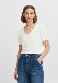 Haut blanc à manches courtes avec un col en V et un tissu texturé, présentant une bordure décorative le long du col et des manches, associé à un jean bleu taille haute.