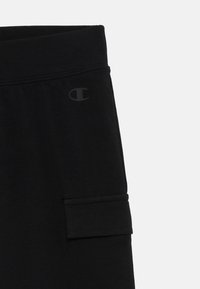 Gros plan sur un pantalon de survêtement noir avec une taille élastique, une poche cargo sur la jambe, et un logo Champion subtil près de la hanche.