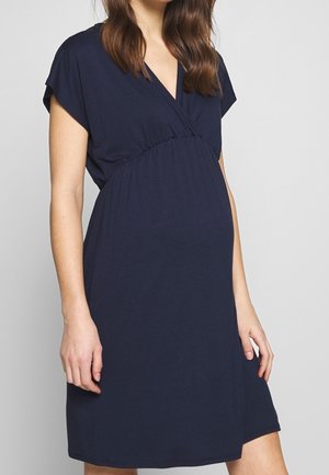 Raskaana oleva nainen pukeutuneena lyhythihaiseen laivastonsiniseen äitiysmekkoon, jossa on V-aukko ja korostettu empire-mallinen vyötärö.