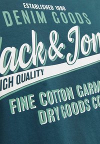 Teal bomulds T-shirt med hvid og grøn tekst: "Etableret 1990", "Denimvarer", "Jack & Jones", "Fine bomuldsbeklædning".