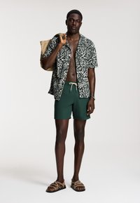 Camicia a maniche corte da uomo con motivo di foglie verdi e bianche, abbinata a pantaloni da bagno verde scuro, con una borsa tote beige e sandali marroni.