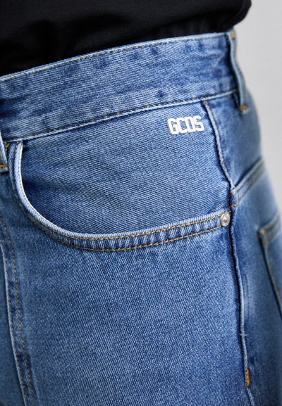 Zilā džinsā ar piegulošu dizainu, oranžiem šuvumiem, redzamu kabatu un baltu "GCDS" logotipu aizmugurē.