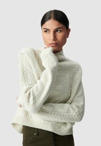 Maglione lavorato a maglia color crema con dettagli a treccia sulle maniche, colletto alto e orlo a coste; texture morbida con leggere variazioni di colore.