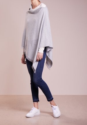 Kobieta w jasnoszarym ponczo z frędzlami na białej koszuli z długim rękawem, w ciemnoniebieskich dżinsach i białych trampkach, stojąca bokiem.