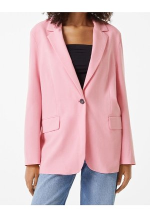 Femme portant un blazer rose à un seul bouton sur un haut noir et un jean bleu, debout, les bras détendus le long du corps.
