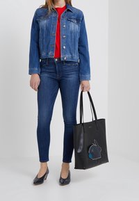 Bolso tote negro con un diseño de conejo y acentos azules. Modelo vistiendo una chaqueta de mezclilla, camiseta roja y jeans azules, con zapatos azul marino.