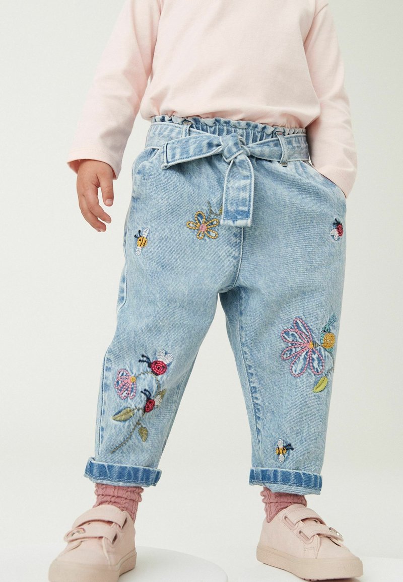 Tout-petit portant un jean bleu clair brodé de fleurs, abeilles, papillons, un haut manche longue rose clair, des chaussettes roses et des chaussures rose pâle.