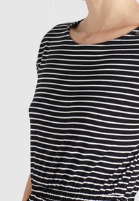 Kvinna som bär en svart- och vitrandig topp med rund halsringning och samlad midja mot en enfärgad bakgrund.