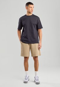 T-shirt in cotone grigio scuro con scollatura a girocollo, abbinato a pantaloni beige. Il modello indossa sneakers bianche con dettagli grigi e calze bianche.