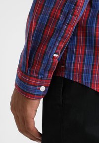 Röd och blå rutigt skjortärm med vita knappar, som har en blandning av vertikala och horisontella ränder, nära vy av manschetten.