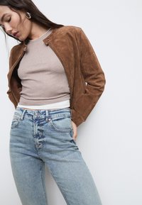 Giacca bomber in suede marrone con colletto rotondo, abbinata a un top a coste beige chiaro e jeans chiari a vita media. Design semplice, senza motivi.