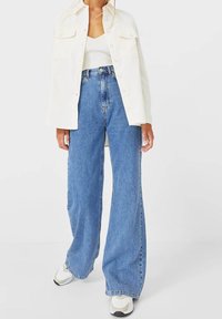 Kobieta w szerokich niebieskich dżinsach, białej, prążkowej koszulce bez rękawów, off-white koszuli zapinanej na guziki oraz białych sneakersach, stojąca na jednolitym tle.