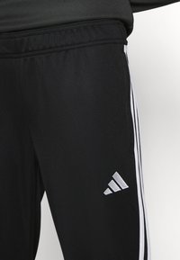Čierne športové nohavice s bielymi pruhmi a logom adidas na dolnej ľavej nohe. Vyrobené z ľahkej, textúrovanej látky.