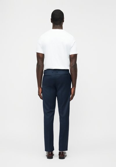 Άνδρας που στέκεται με την πλάτη στραμμένη, φοράει λευκό πουκάμισο με κοντά μανίκια, σκούρο μπλε παντελόνι φόρμας και σκούρα καφέ επίσημα παπούτσια, σε απλό φωτεινό φόντο.