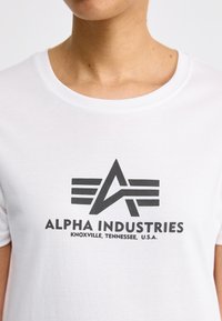 Valkoinen puuvillapaita, jossa on musta graafinen logo, jossa on tähti ja vaakasuorat viivat. Kaula-aukko on pyöreä ja kangas näyttää sileältä.