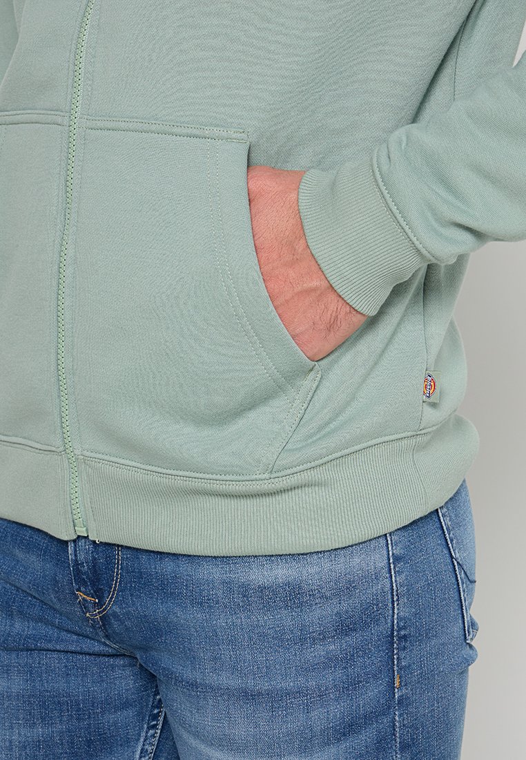 Sudadera con cremallera de color verde claro con un acabado texturizado, presenta bolsillos delanteros y puños acanalados. Combinada con unos pantalones vaqueros de mezclilla azul.