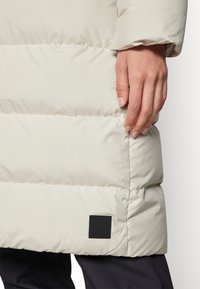 Puffjacka i ljusbeige med sydd paneldesign. Har en svart logotyp-patch och en slät, glänsande yta. Synlig hand som greppar fållen.