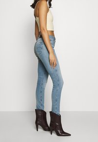 Mujer vestida con jeans ajustados de color azul claro, top corto beige y botines de tacón alto negros con punta afilada, de pie de lado.