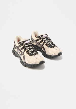 Paire de baskets de sport beige et noires avec dessus en mesh et synthétique, semelles noires, et lacets élastiques sans nœud avec boutons roses.