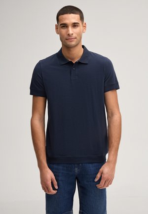 Man met kort haar die een marineblauw poloshirt met korte mouwen en een blauwe spijkerbroek draagt, staande tegen een effen lichte achtergrond.