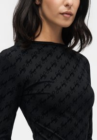 Femme portant un haut noir à manches longues avec un motif subtil en écriture répétée et un col rond, raccourci au niveau du menton et de la poitrine.