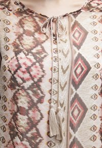 Top de tela ligera con un estampado floral y geométrico en tonos de rosa, beige y marrón. Presenta un escote de ojal y acentos de borlas.