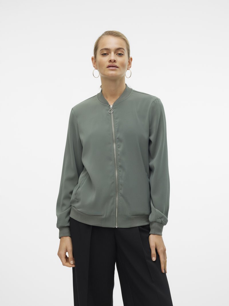 Femme portant une veste bomber verte zippée et un pantalon noir, debout devant un fond blanc uni.