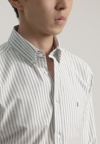 Camisa de botones de rayas en blanco con franjas verticales grises, con un cuello puntiagudo y un pequeño detalle de logo en el pecho. Textura suave.