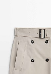 Jupe beige avec des boutons noirs croisés, une ceinture assortie avec une boucle noire rectangulaire et une poche latérale verticale.
